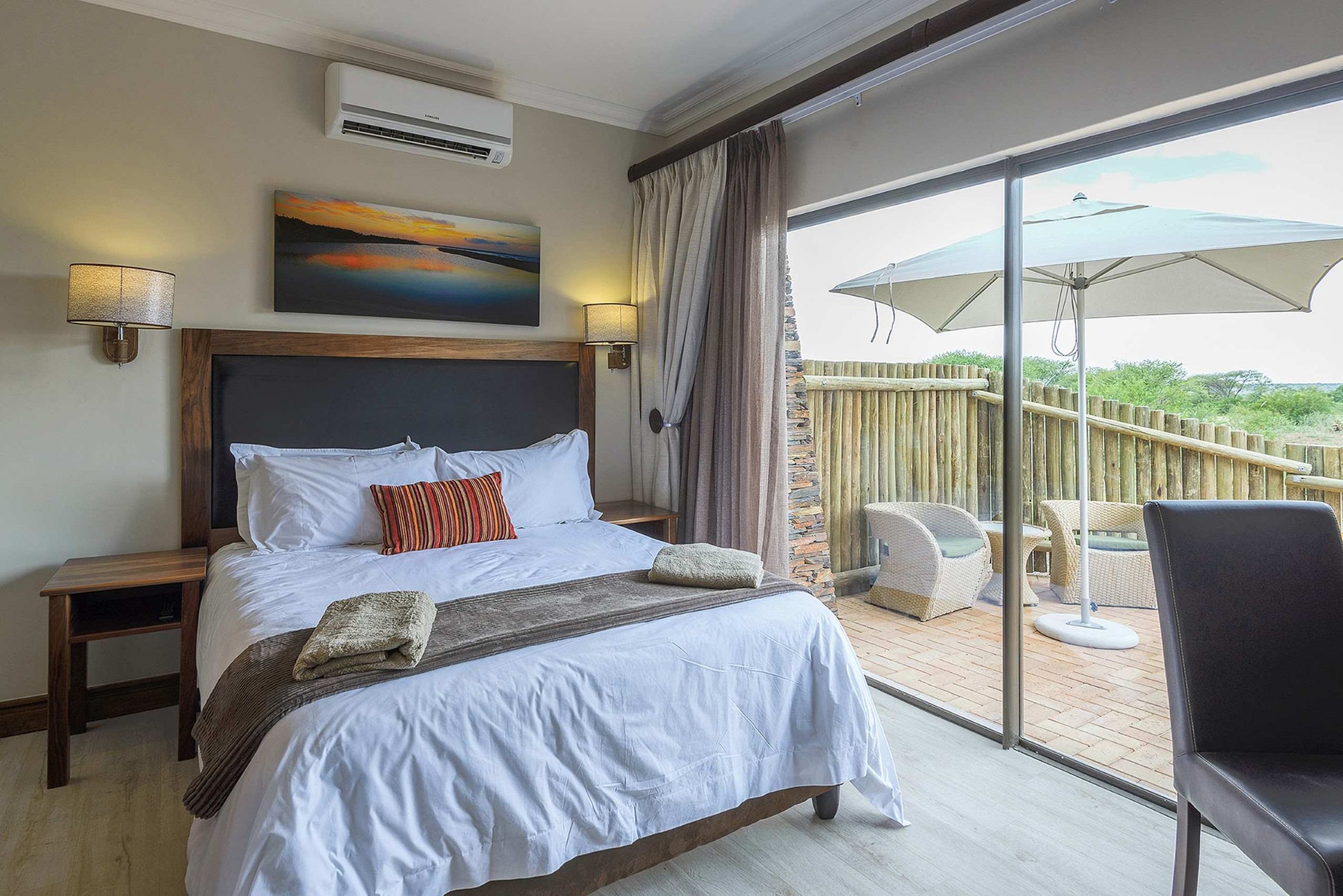 A modern bedroom interior with a neatly made bed, wooden headboard, wall-mounted lamps, and large sliding glass doors opening to a balcony with patio furniture and an umbrella.